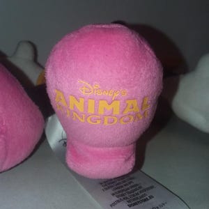 May include: A pink plush toy with the text "Disney's Animal Kingdom" in yellow. The toy has a rounded top and a cylindrical base. Other plush toys are partially visible in the background.
