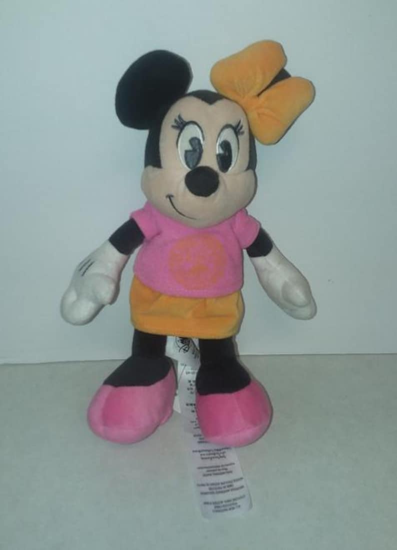 May include: A plush Minnie Mouse doll with a pink shirt, orange skirt, and a yellow bow. The doll has black ears, a black nose, and pink shoes. The doll is standing upright.