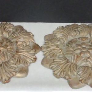 May include: Two decorative wall plaques in the shape of a flower with a raised center. The plaques are made of a light brown material with a gold finish.