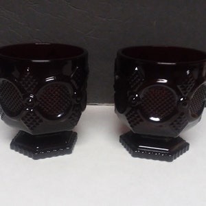 May include: Two dark red glass goblets with a textured pattern. The goblets have a square base and a detailed design around the body. The glass has a glossy finish, and the goblets are likely vintage or antique.