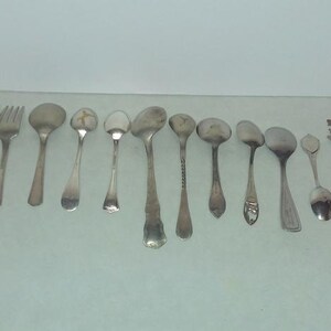 May include: A collection of 12 silver spoons and forks, including a fork with a decorative handle. The spoons are various sizes and styles, some with ornate designs.