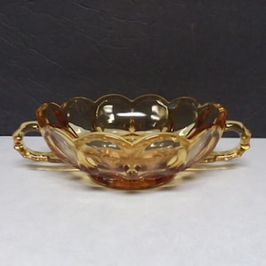 May include: A translucent amber-colored glass bowl featuring a scalloped rim and two side handles. The exterior surface of the bowl displays a molded pattern with rounded indentations and a circular base, reflecting light across its smooth surface.