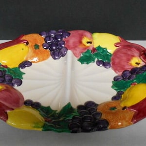 May include: A white ceramic divided serving dish with a fruit design. The dish is decorated with red, yellow, green, and purple fruit, and has two red bows on the sides.