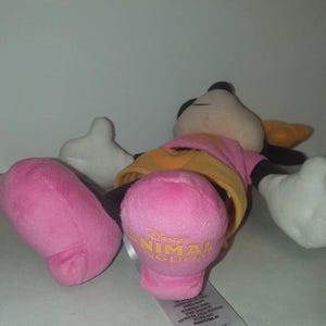 May include: A plush Minnie Mouse doll, lying on its back. The doll features a pink dress, yellow undershirt, and pink feet with "Disney's Animal Kingdom" printed in gold. The doll has black ears, a black nose, and white gloves.