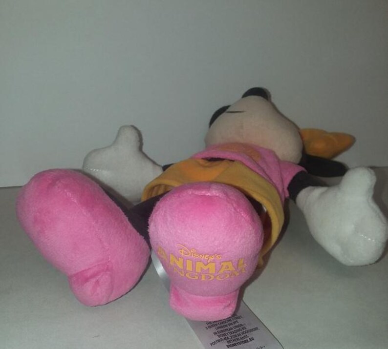 May include: A plush Minnie Mouse doll, lying on its back. The doll features a pink dress, yellow undershirt, and pink feet with "Disney's Animal Kingdom" printed in gold. The doll has black ears, a black nose, and white gloves.