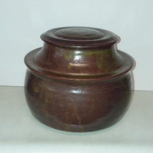 May include: A handcrafted, glazed ceramic jar with a lid. The jar is a deep reddish-brown color with subtle green and gold highlights. The lid has a rounded top and fits snugly on the jar. The jar has a rounded body and a wide, flat base. This decorative jar is suitable for storage.