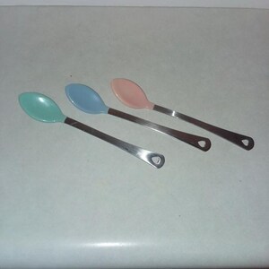 May include: Three spoons with pastel-colored bowls and stainless steel handles. The spoon bowls are light green, blue, and pink. The handles have a heart-shaped cutout at the end. Kitchen utensils.