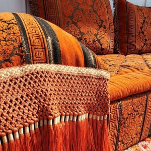 May include: A brown and orange couch with a patterned fabric. The couch has a fringe trim and a crocheted detail. The couch is in a living room setting.