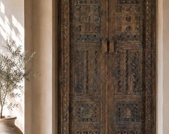 May include: Ornate, dark brown wooden double doors with detailed carvings. The doors have a central handle and are set within a neutral-toned wall. The overall design suggests a traditional or antique style.