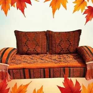 May include: A traditional Moroccan sofa with two cushions, featuring a rich orange and black patterned fabric. The sofa is decorated with tassels and fringe. The background features a blurred image of autumn leaves.