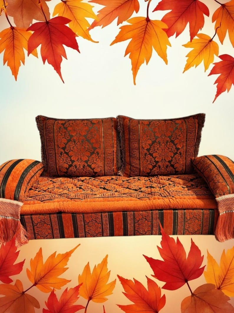 May include: A traditional Moroccan sofa with two cushions, featuring a rich orange and black patterned fabric. The sofa is decorated with tassels and fringe. The background features a blurred image of autumn leaves.