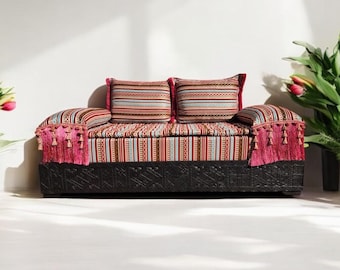 May include: A traditional Moroccan sofa with a black wooden frame and colorful striped upholstery. The sofa has two cushions with a geometric pattern and pink tassels.