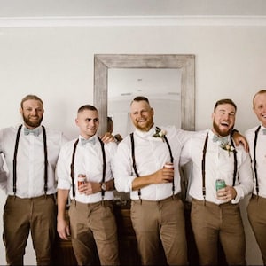 Puede incluir: Cinco hombres con camisas blancas, pantalones marrones y tirantes. Todos sonríen y sostienen latas de cerveza. Los hombres están de pie frente a un espejo con un marco de madera.
