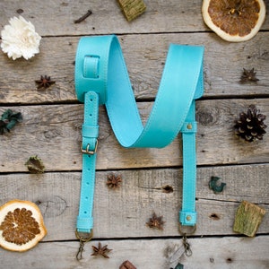 May include: A turquoise leather guitar strap with adjustable length. The strap features brass-colored hardware, including buckles and swivel hooks. The strap is shown on a rustic wooden surface with decorative elements like dried fruit and star anise.
