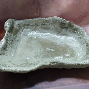 May include: A handmade, irregularly shaped, shallow bowl with a textured, off-white surface. The bowl has a rough, uneven rim and appears to be made of a composite material. The interior has a slightly glossy finish.