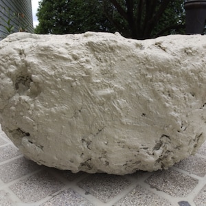 May include: A large, light-colored, textured stone bowl with a rough, uneven surface. The bowl has a wide, open top and sits on a tiled surface. The neutral color and organic shape suggest a rustic or natural aesthetic.