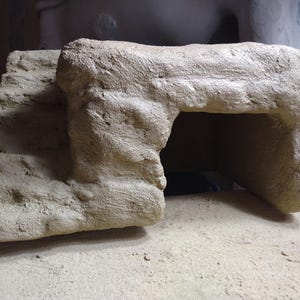 May include: A beige, textured, cave-like structure with an arched opening. The structure has a rough, stone-like appearance and sits on a flat, light-colored surface. The overall design suggests a miniature habitat or decorative element.