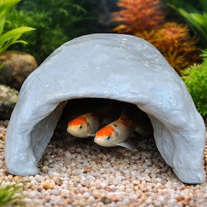 May include: Two orange and white koi fish swim under a light gray, cave-shaped aquarium decoration. The fish are in a tank with gravel and surrounded by green and orange aquatic plants. The scene is well-lit and detailed.