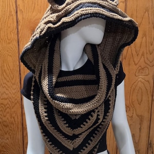 Op de afbeelding: Een gehaakte capuchon-cowl met een gestreept patroon in beige en zwart, voorzien van een brede, uitlopende capuchon en een puntig, driehoekig voorpand. De capuchon heeft twee kleine, afgeronde, oorvormige uitsteeksels van bijpassend beige garen.
