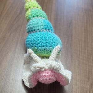 May include: A crocheted snail toy featuring a gradient shell in shades of green and blue. The snail's body is white with pink and white details, including the face and antennae. The toy is crafted from soft yarn.