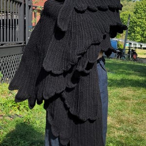 May include: A black knitted shawl with a scalloped edge design. The shawl is worn over the shoulders and drapes down the front. The garment is made of a thick, textured yarn.