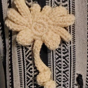 May include: A cream-colored, crocheted flower with multiple petals and a stem. The flower is lying on a black and white patterned fabric. A small, black raven pin is also visible.