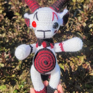 Puede incluir: Muñeca de ganchillo hecha a mano con cuerpo blanco, detalles en rojo y negro y campanas doradas. La muñeca tiene cuernos, un diseño en espiral en el vientre y se sostiene frente a un fondo verde borroso. El diseño de la muñeca recuerda a un bufón.