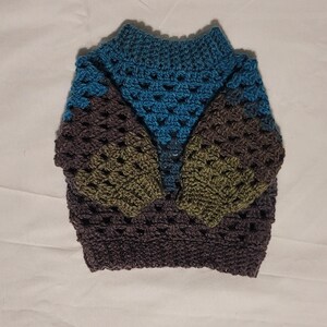 Könnte beinhalten: Ein handgefertigter Häkelpullover in Blau-, Grün- und Brauntönen. Der Pullover hat einen hohen Kragen und lange Ärmel. Das Häkelmuster erzeugt ein strukturiertes Aussehen.