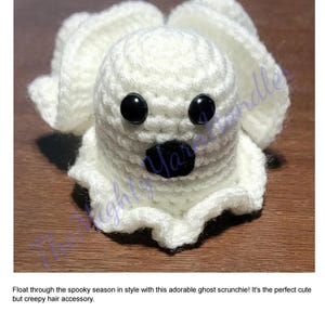 Op de afbeelding: Een handgemaakte witte gehaakte spook scrunchie met zwarte knoopogen en een zwarte mond. De scrunchie is een haaraccessoire met Halloween-thema. De tekst "Ghost Scrunchie Crochet Pattern" staat bovenaan.