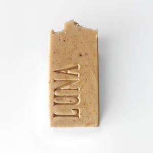May include: A light brown rectangular soap bar with the word "LUNA" imprinted on it. The soap has a textured surface with small flecks visible. A small section of the top edge is missing, indicating use. The soap is on a white background.