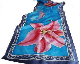 May include: A blue silk scarf with a pink lily flower design. The scarf has a navy blue border with a white decorative pattern.