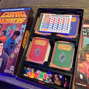 May include: A board game called "Comic Hunters" with a Marvel logo. The game box features colorful illustrations of comic book characters. The game includes a score sheet, game cards, and tokens.