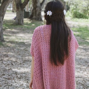 Puede incluir: Un top de crochet rosa con un ajuste holgado y fluido. El top tiene un dise&ntilde;o simple de punto abierto y lo lleva una persona con el pelo largo y oscuro. Dos pinzas para el pelo en forma de margarita blanca est&aacute;n en el pelo de la persona.