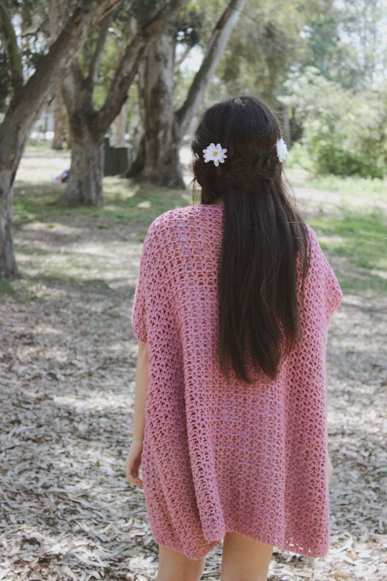 Puede incluir: Un top de crochet rosa con un ajuste holgado y fluido. El top tiene un dise&ntilde;o simple de punto abierto y lo lleva una persona con el pelo largo y oscuro. Dos pinzas para el pelo en forma de margarita blanca est&aacute;n en el pelo de la persona.