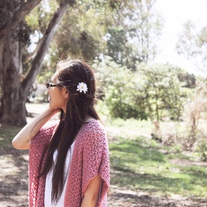 Puede incluir: Una mujer con el pelo largo y casta&ntilde;o lleva un c&aacute;rdigan de ganchillo rosa con una flor blanca en el pelo. Est&aacute; de pie en un parque con &aacute;rboles y hierba de fondo.