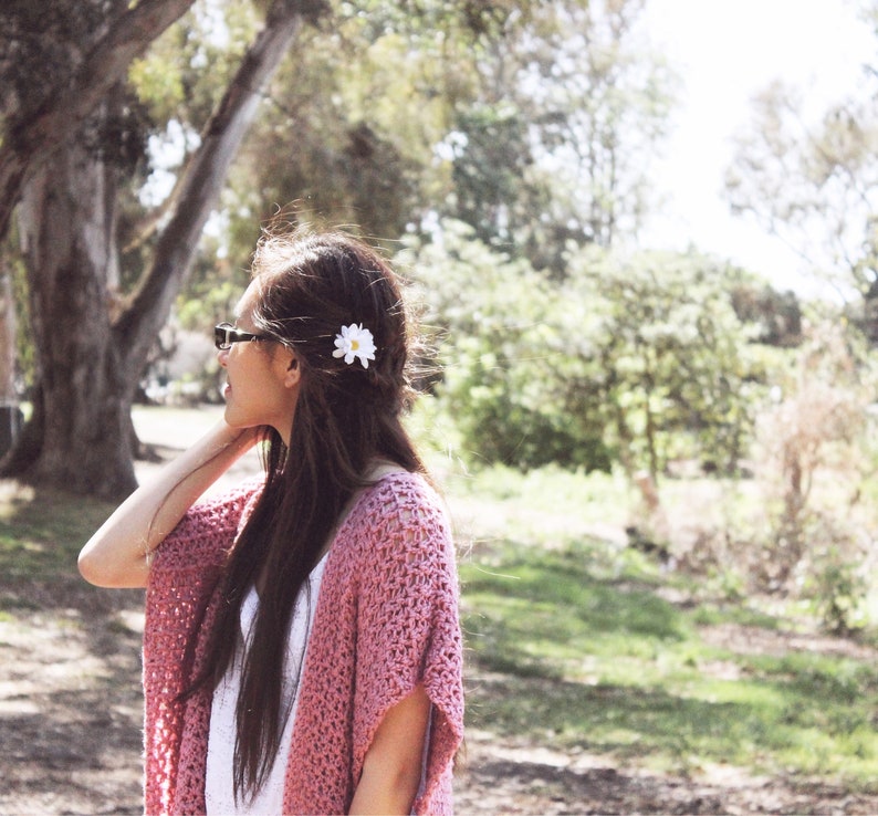 Puede incluir: Una mujer con el pelo largo y casta&ntilde;o lleva un c&aacute;rdigan de ganchillo rosa con una flor blanca en el pelo. Est&aacute; de pie en un parque con &aacute;rboles y hierba de fondo.