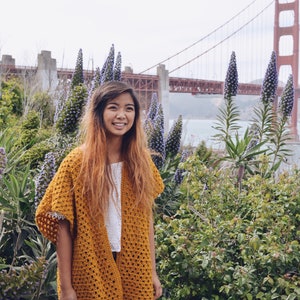 Puede incluir: Una mujer con un c&aacute;rdigan de crochet amarillo mostaza, una blusa blanca y pantalones negros, de pie frente a un campo de flores moradas con el puente Golden Gate al fondo.