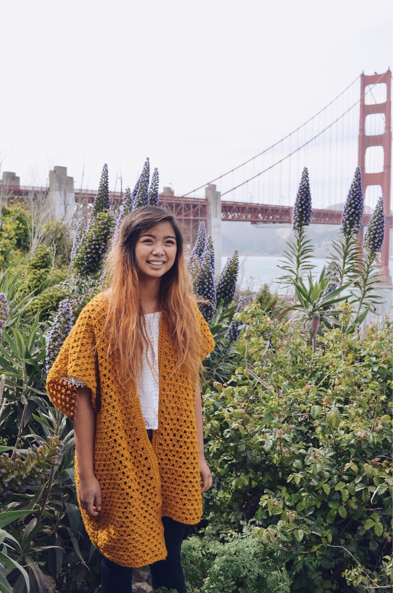 Puede incluir: Una mujer con un c&aacute;rdigan de crochet amarillo mostaza, una blusa blanca y pantalones negros, de pie frente a un campo de flores moradas con el puente Golden Gate al fondo.