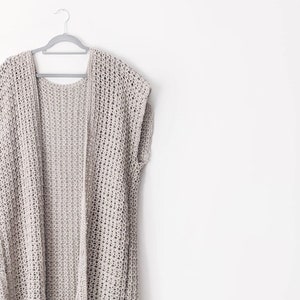 Puede incluir: Un c&aacute;rdigan de crochet gris claro de corte holgado y sin mangas, colgado de una percha plateada sobre un fondo blanco.
