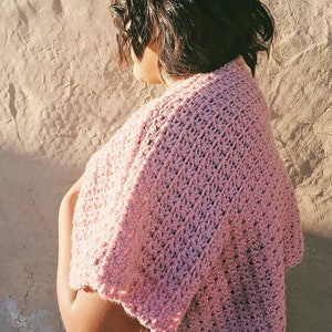Puede incluir: Una estola de ganchillo rosa con un delicado patr&oacute;n de puntadas. La estola la lleva puesta una persona con el pelo oscuro, pero el foco est&aacute; en la prenda.