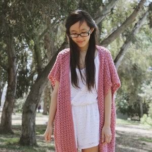 Puede incluir: Un c&aacute;rdigan de crochet rosa con mangas cortas y abertura frontal. El c&aacute;rdigan se lleva sobre un vestido blanco.