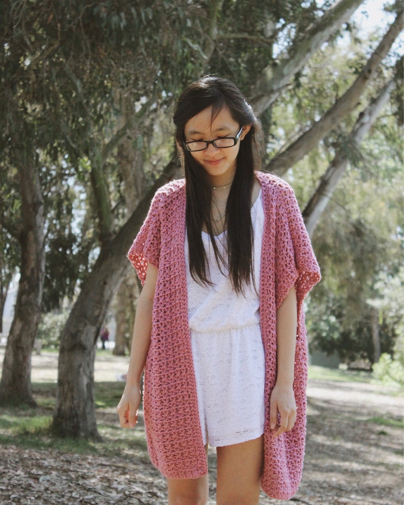 Puede incluir: Un c&aacute;rdigan de crochet rosa con mangas cortas y abertura frontal. El c&aacute;rdigan se lleva sobre un vestido blanco.