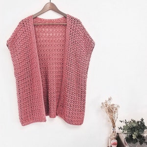 Puede incluir: Un c&aacute;rdigan de crochet rosa polvoriento con un corte holgado y mangas cortas. El c&aacute;rdigan est&aacute; colgado en una percha de madera contra una pared blanca.