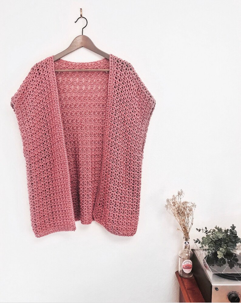 Puede incluir: Un c&aacute;rdigan de crochet rosa polvoriento con un corte holgado y mangas cortas. El c&aacute;rdigan est&aacute; colgado en una percha de madera contra una pared blanca.