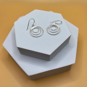 May include: A pair of silver earrings with a circular design of three concentric rings. The earrings are presented on a white geometric surface against a light orange backdrop. The earrings have a hook closure.