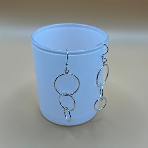 May include: Silver-coloured dangle earrings with a series of connected circular hoops. The earrings hang from a simple hook and are shown against a soft, neutral background. The design is minimalist and modern, suitable for everyday wear.
