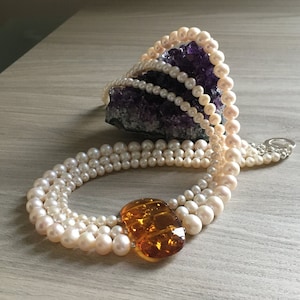 May include: A multi-strand pearl necklace with a large amber pendant. The necklace is made of white pearls and the pendant is a rich, warm amber colour.