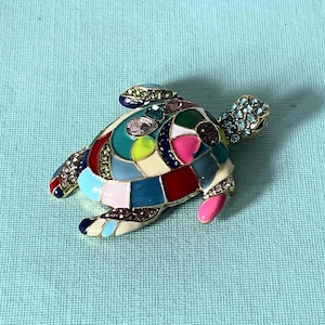 Pode incluir: Um broche em forma de tartaruga com uma estrutura de metal dourado preenchida com seções de esmalte multicoloridas em tons de vermelho, azul, rosa, amarelo e branco. O casco e as nadadeiras são acentuados com pequenos strass brilhantes.
