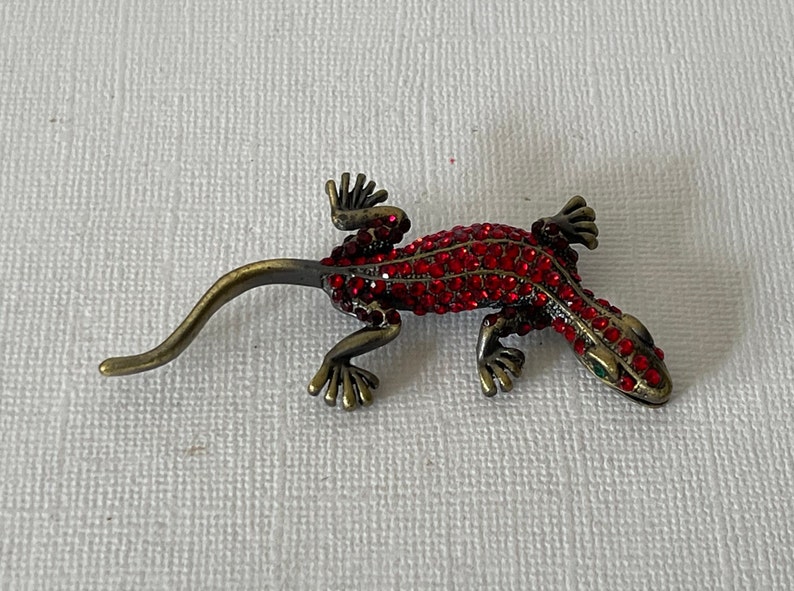 May include: A red jeweled gecko brooch with green eyes. The gecko is made of metal and has a long tail.