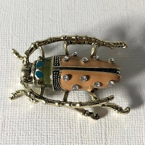 May include: A gold-toned beetle brooch featuring a peach-colored body with embedded clear rhinestones. The insect has a green head section with two bright blue accents and textured gold legs extending outward from the central body structure.